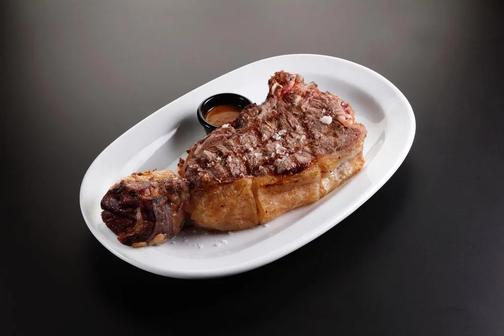 A thick bone-in grilled chuleton, crusted and flecked with salt flakes, on a white oval platter with a small pot of sauce, against a dark studio background