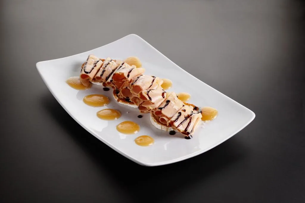 Rolled slices of pale cured meat over rounds of white cheese along a rectangular white plate, zigzagged with dark balsamic reduction and dotted with amber sauce