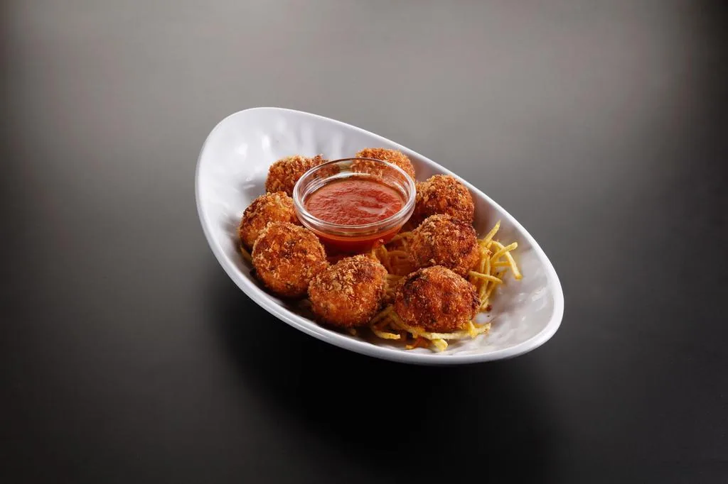 Eight breadcrumbed fried balls around a small glass pot of red sauce, on a bed of straw potatoes in a curved white dish on a dark background