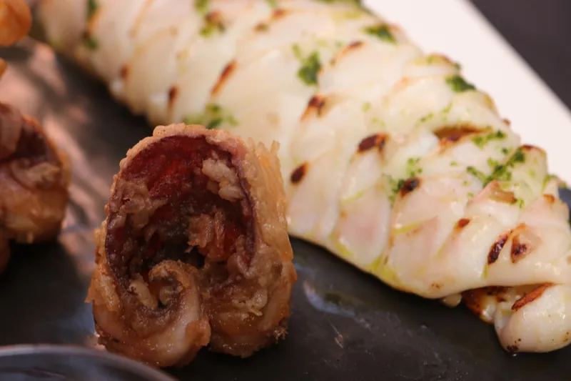 A whole grilled squid, scored in a diamond pattern and scattered with chopped parsley and oil, beside a browned stuffed piece, on a dark slate