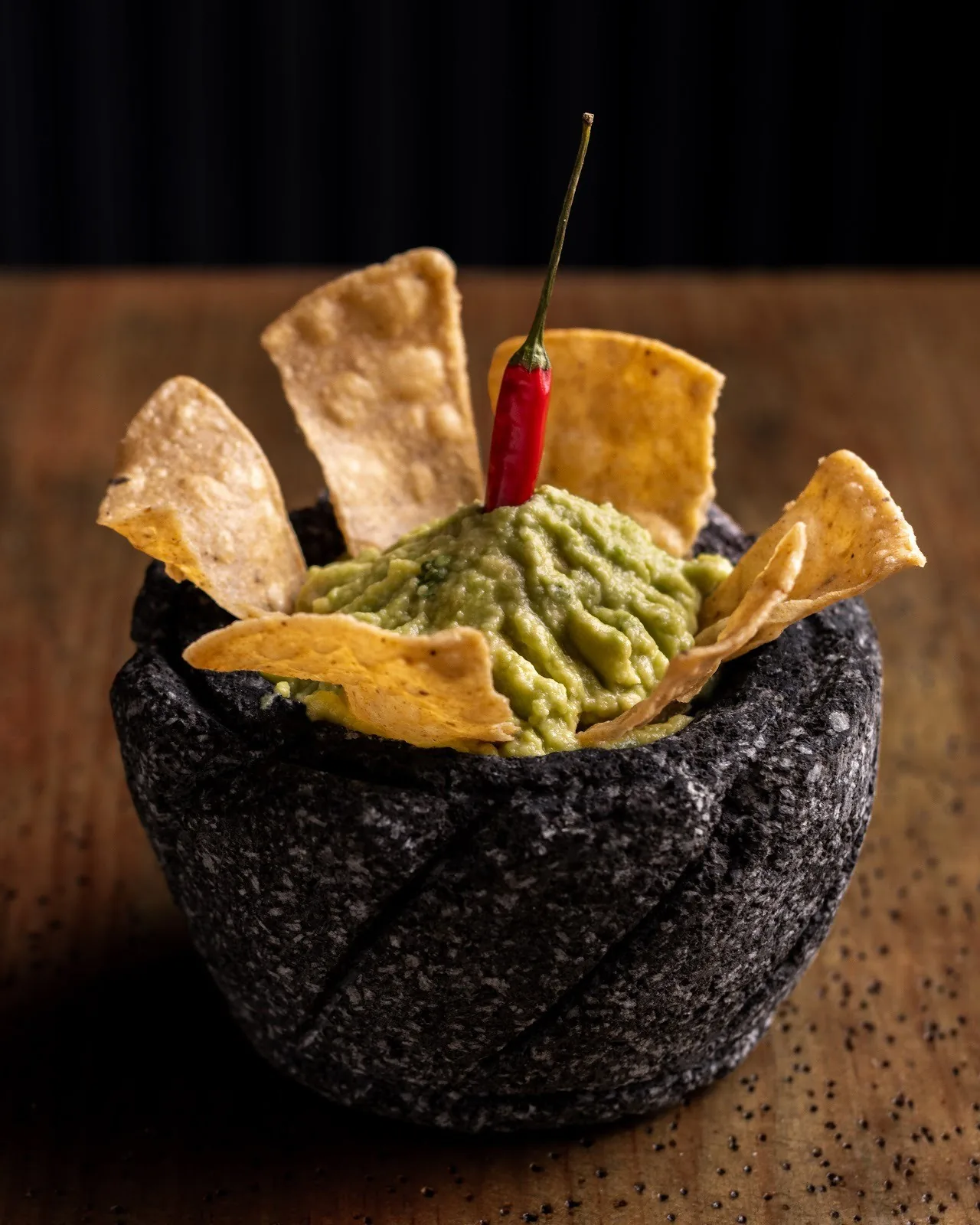 A stone bowl of guacamole topped with tortilla chips and a small red chilli