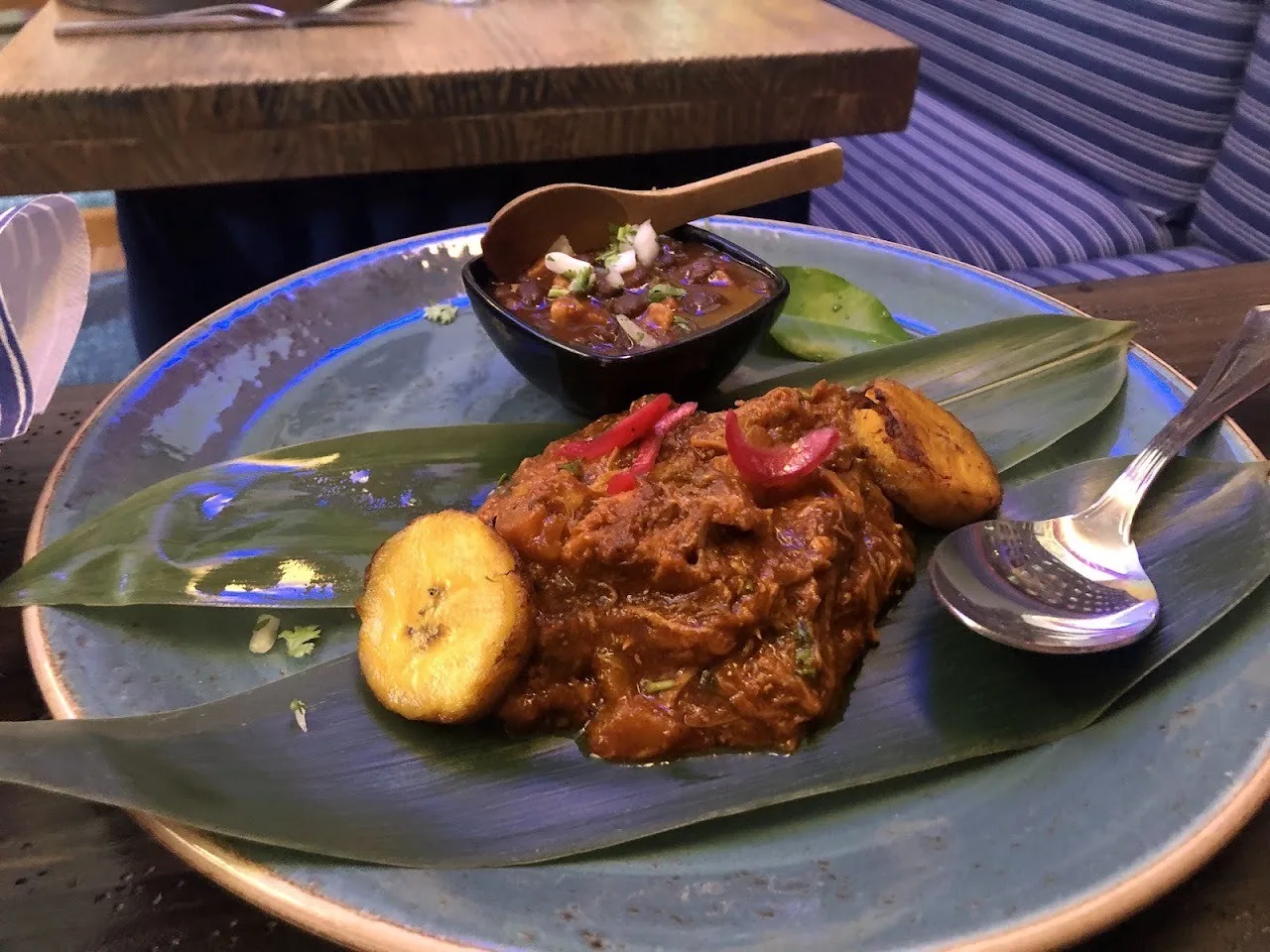 A grey plate of stewed red meat with fried plantain slices, a small bowl of sauce and a lime wedge on a leaf