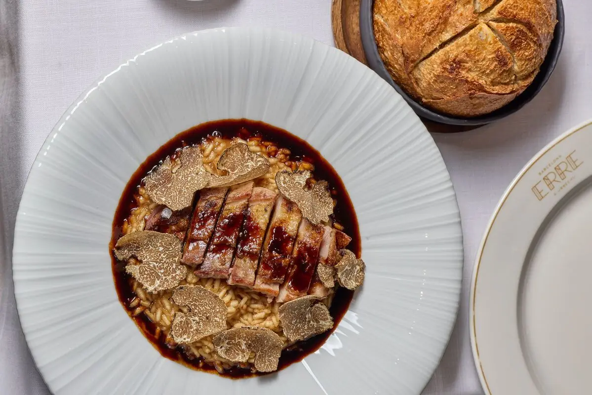 Overhead shot of a white ribbed plate with sliced roasted meat over rice topped with shaved black truffle, served alongside a round crusty loaf of bread, at Erre & Urrechu