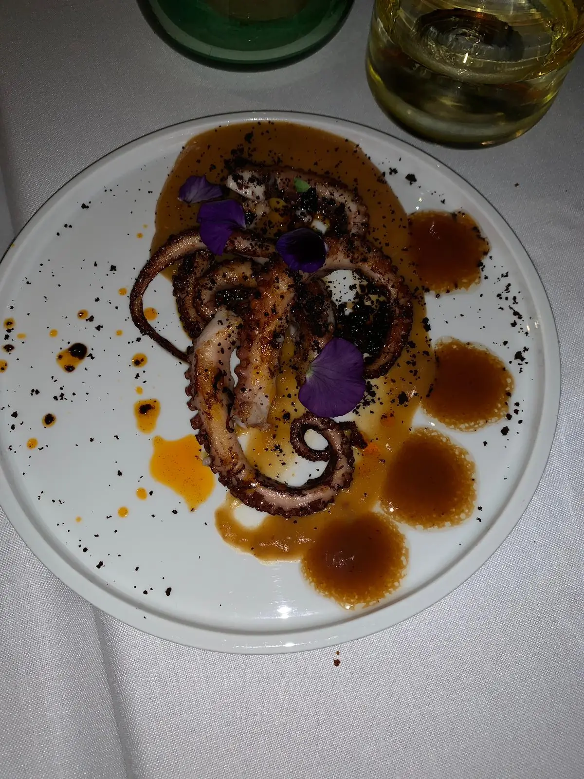 A charred octopus dish plated with purple edible flowers and a dark sauce.