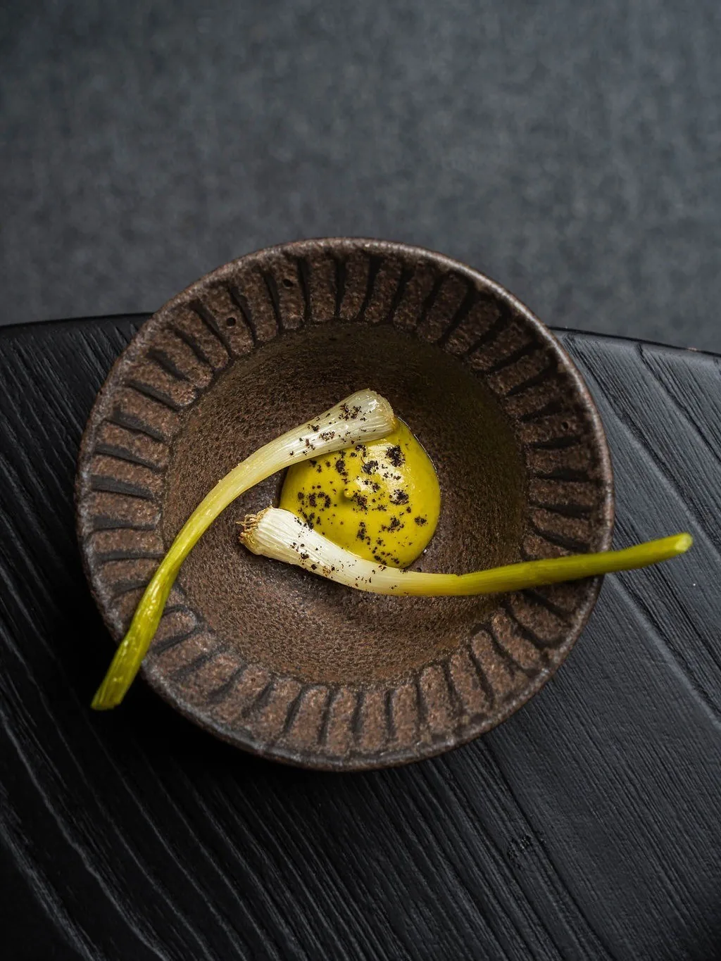 A brown bowl holding yellow sauce and a pale green stalk on a dark table