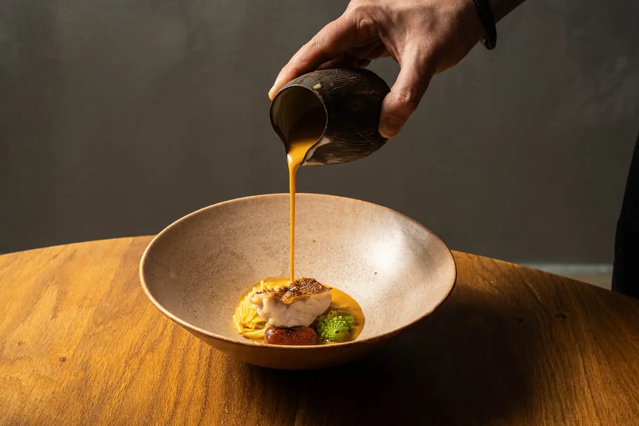 Hand pouring orange sauce over a fish fillet with pasta, chorizo, and romanesco in a ceramic bowl