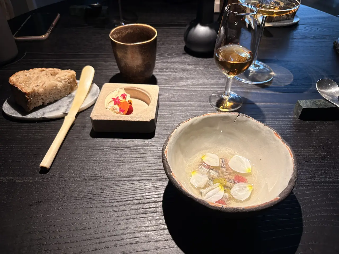Bread with butter, a flower-topped amuse, and a bowl of fish with edible petals beside glasses of wine