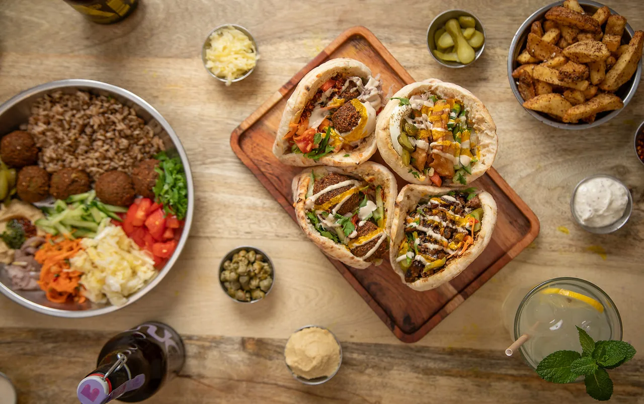 Overhead spread on a pale wood table: four stuffed pitas on a wooden board surrounded by steel bowls of falafel and rice, pickles, spiced chips, hummus, tahini and a lemonade with mint