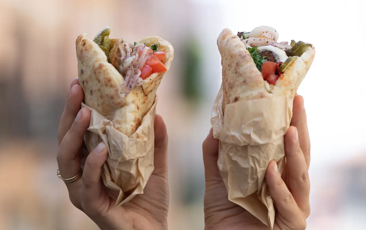 Two hands holding up filled pitas wrapped in crumpled kraft paper, stuffed with falafel, tomato, pickles, sumac onion and white sauce, against a blurred pale street