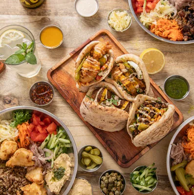 Overhead square crop of three stuffed pitas on a wooden board, ringed by steel bowls of salads, pickles, hummus, capers and a mint lemonade on a pale wood table