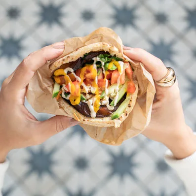Two hands cradling a pita in kraft paper, packed with falafel, cucumber, tomato, mango sauce and tahini, held over the venue's blue star-patterned tiles