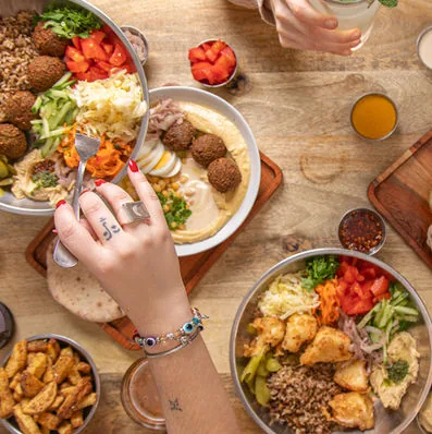 Overhead table of steel bowls holding falafel, hummus, salads and rice, with a hand reaching in with a fork over the hummus bowl