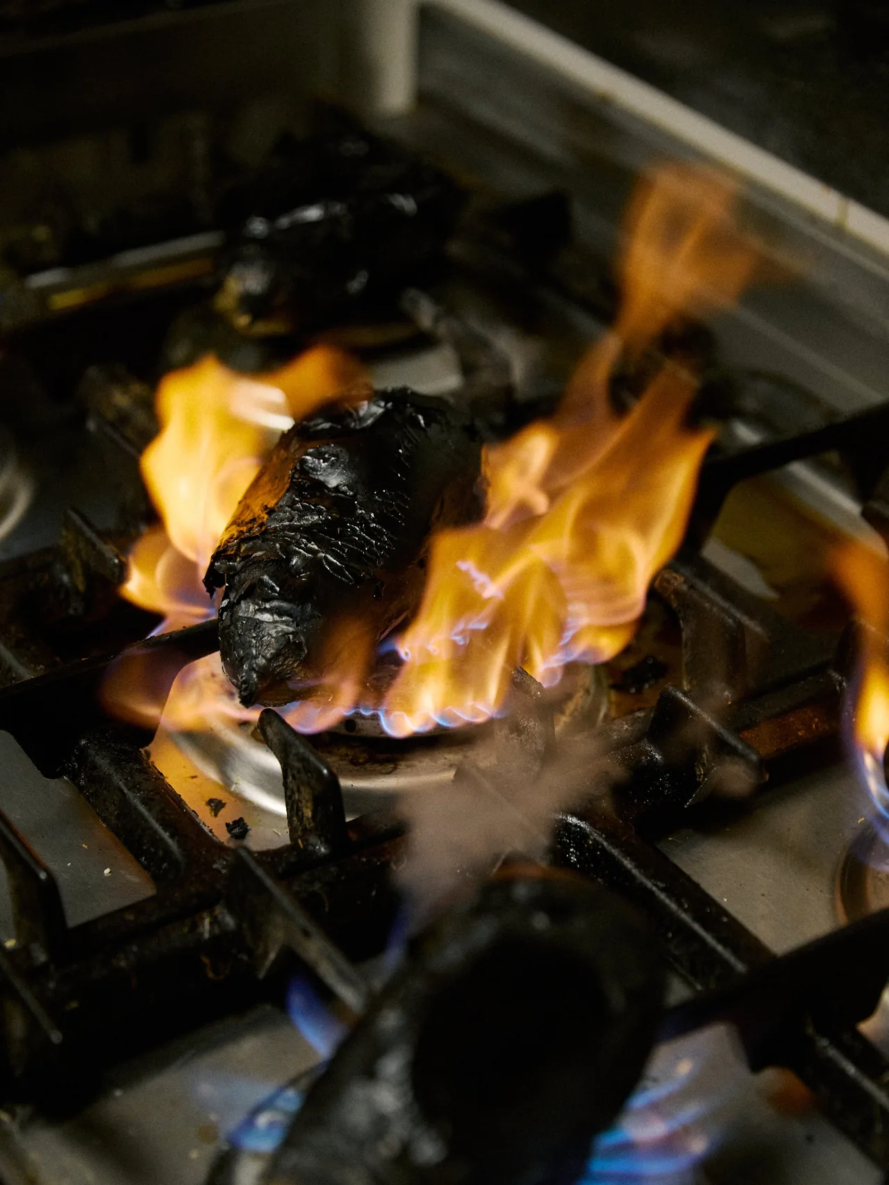 Aubergines charring black directly in the orange flames of a restaurant gas burner, skins blistered and smoking