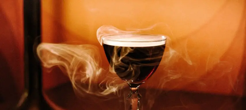 Dark stemmed cocktail wreathed in smoke against a warm amber background