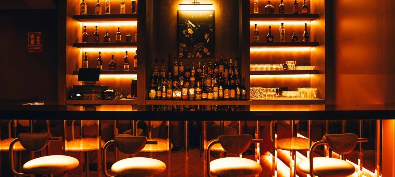 A warmly lit bar with backlit shelves of bottles and a row of stools