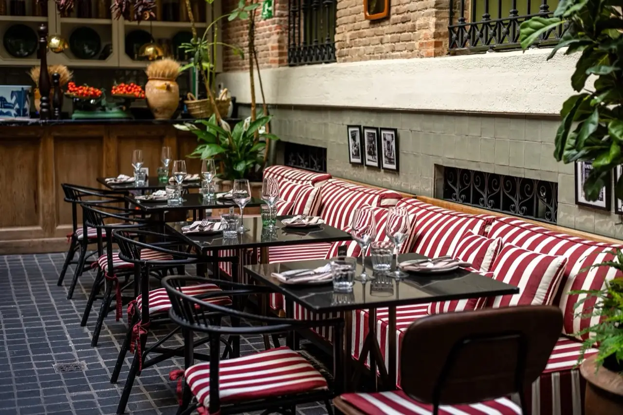 Restaurant courtyard seating with red-and-white striped banquette seating along a tiled wall, black bistro tables set with wine glasses, brick walls and potted plants.
