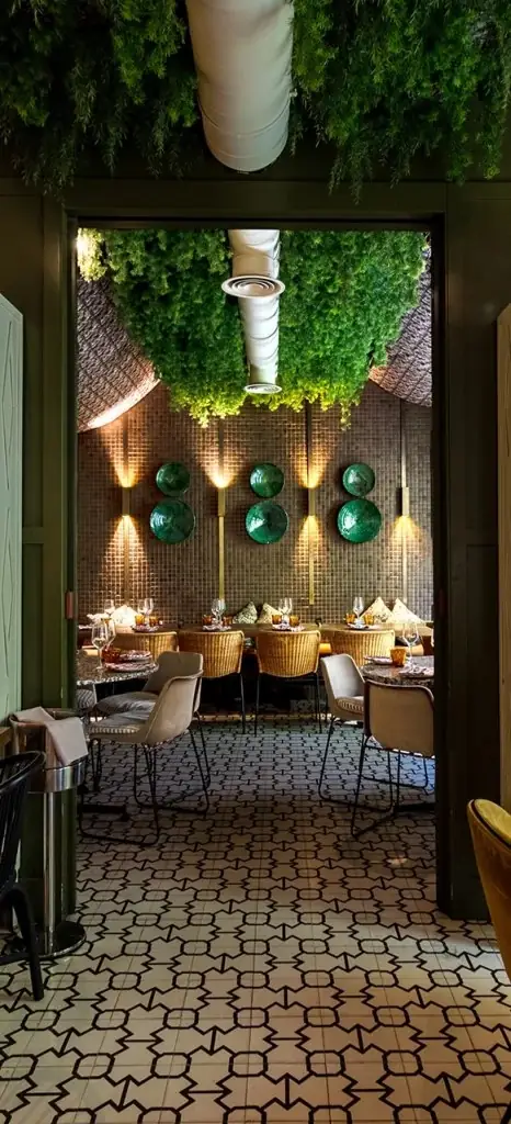 View through a doorway into a dining room with a hanging green plant ceiling installation, black-and-white patterned tile floor, wicker chairs and round green ceramic plates mounted on a tiled wall.