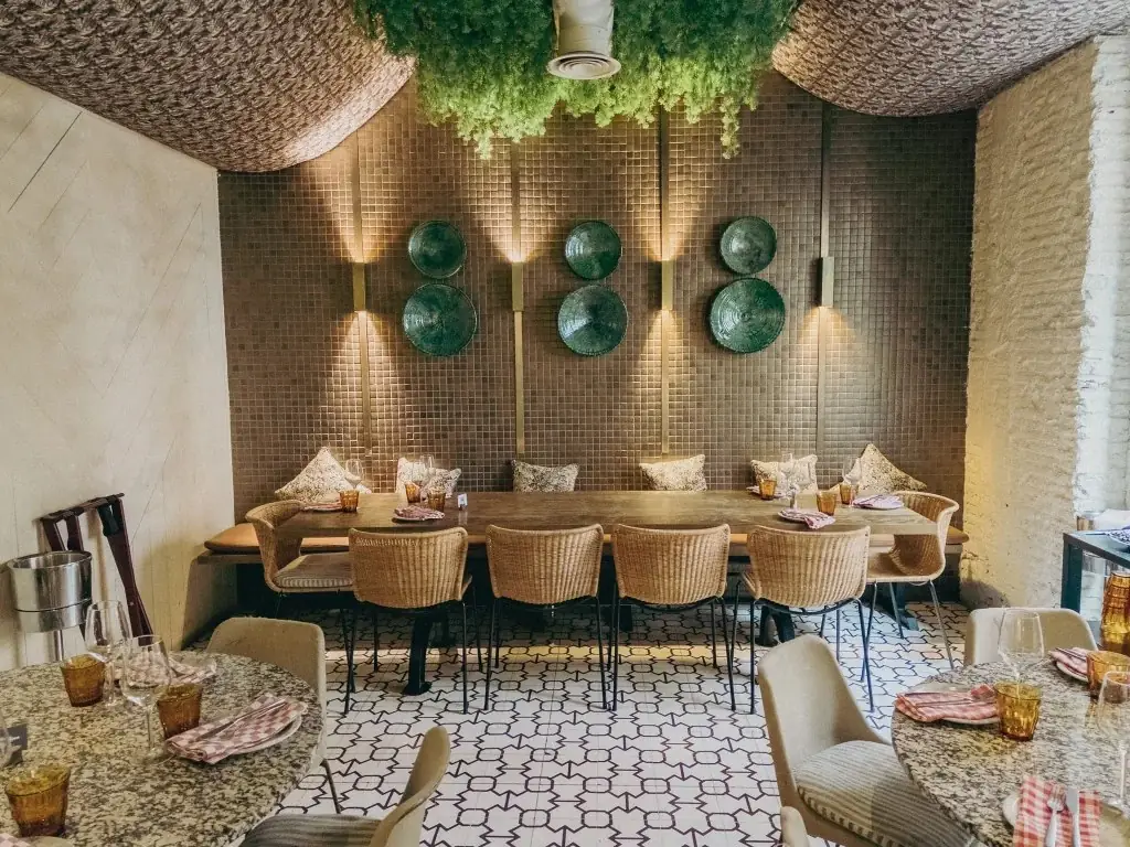 Dining room with a long wooden table surrounded by wicker chairs beneath a hanging greenery ceiling installation, green ceramic plates on a tiled wall, and round tables set with striped napkins in the foreground.