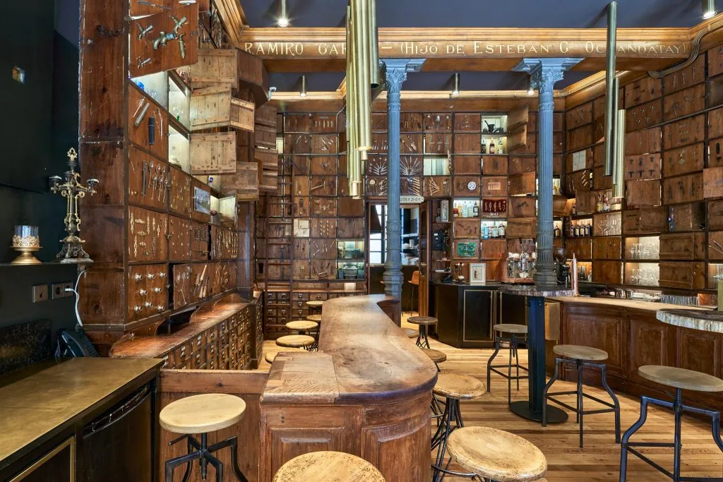 A bar room lined floor to ceiling with the original ironmonger's wooden drawer cabinets and taps, a curved timber counter with stools, and a carved sign reading