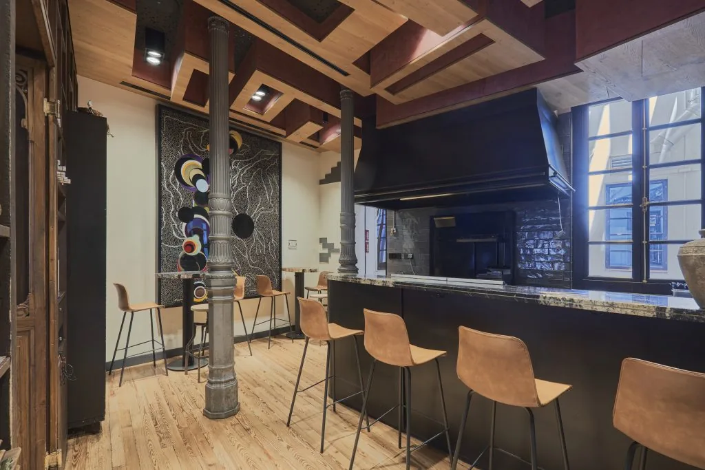 A stone counter with tan leather stools facing an open black-hooded wood-fire grill, under a slatted wood ceiling with cast-iron columns