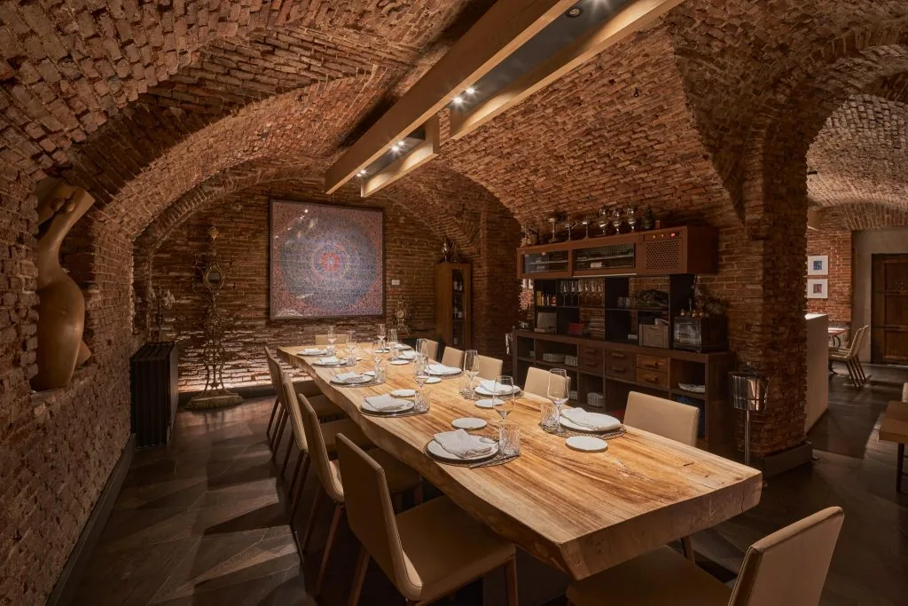 A brick-vaulted cellar dining room with a long live-edge wooden table laid for ten, a large framed artwork and a shelved service station