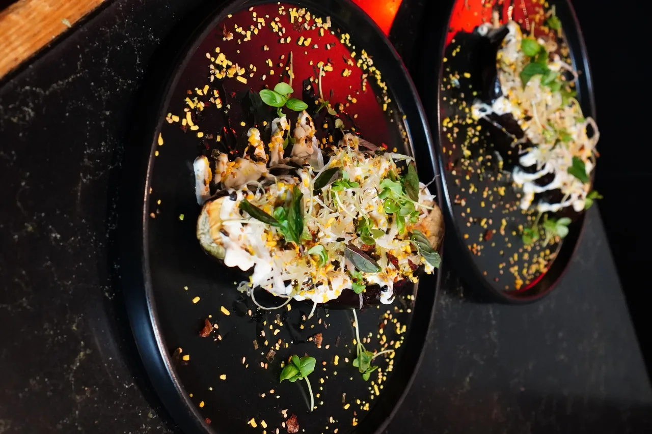 Two black plates with a topped roasted vegetable, white sauce, seeds and small green leaves