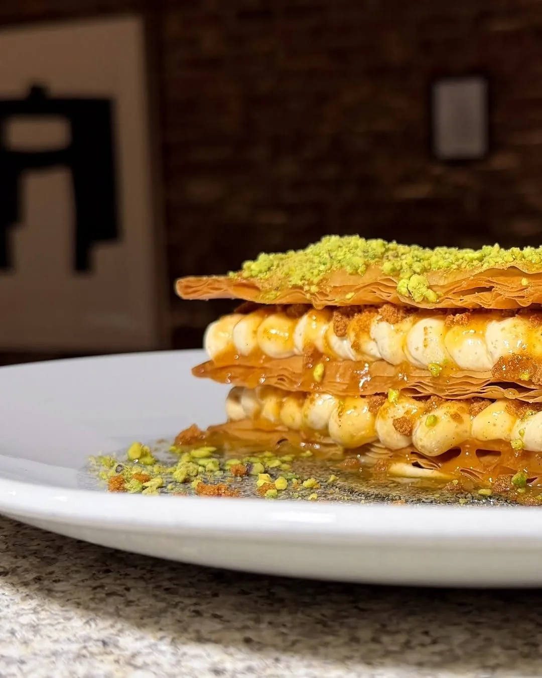 A stack of golden layered pastry with a pale filling and green pistachio topping on a white plate