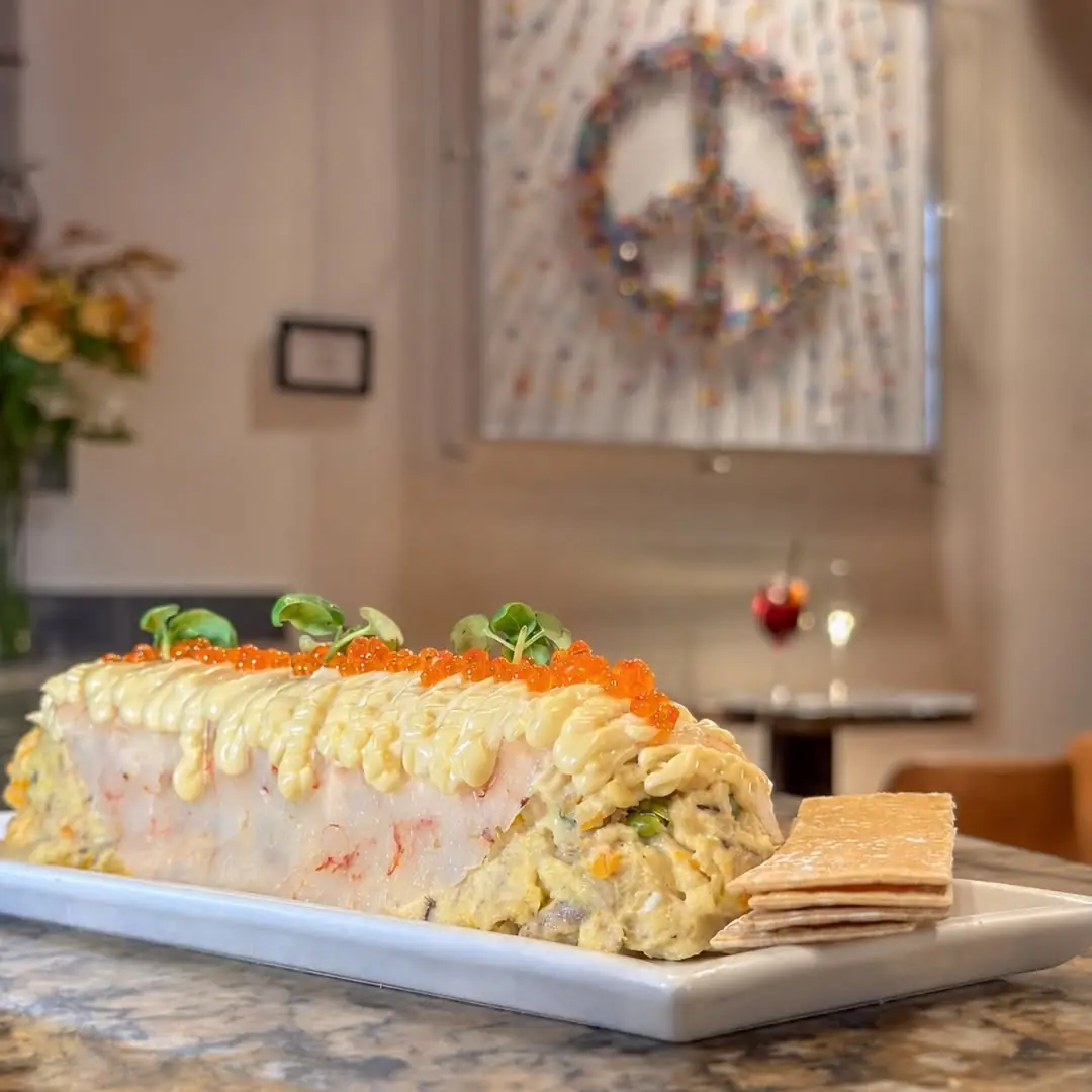 An elongated seafood roll topped with mayonnaise piping, red roe, and microgreens, served with crackers on a rectangular plate