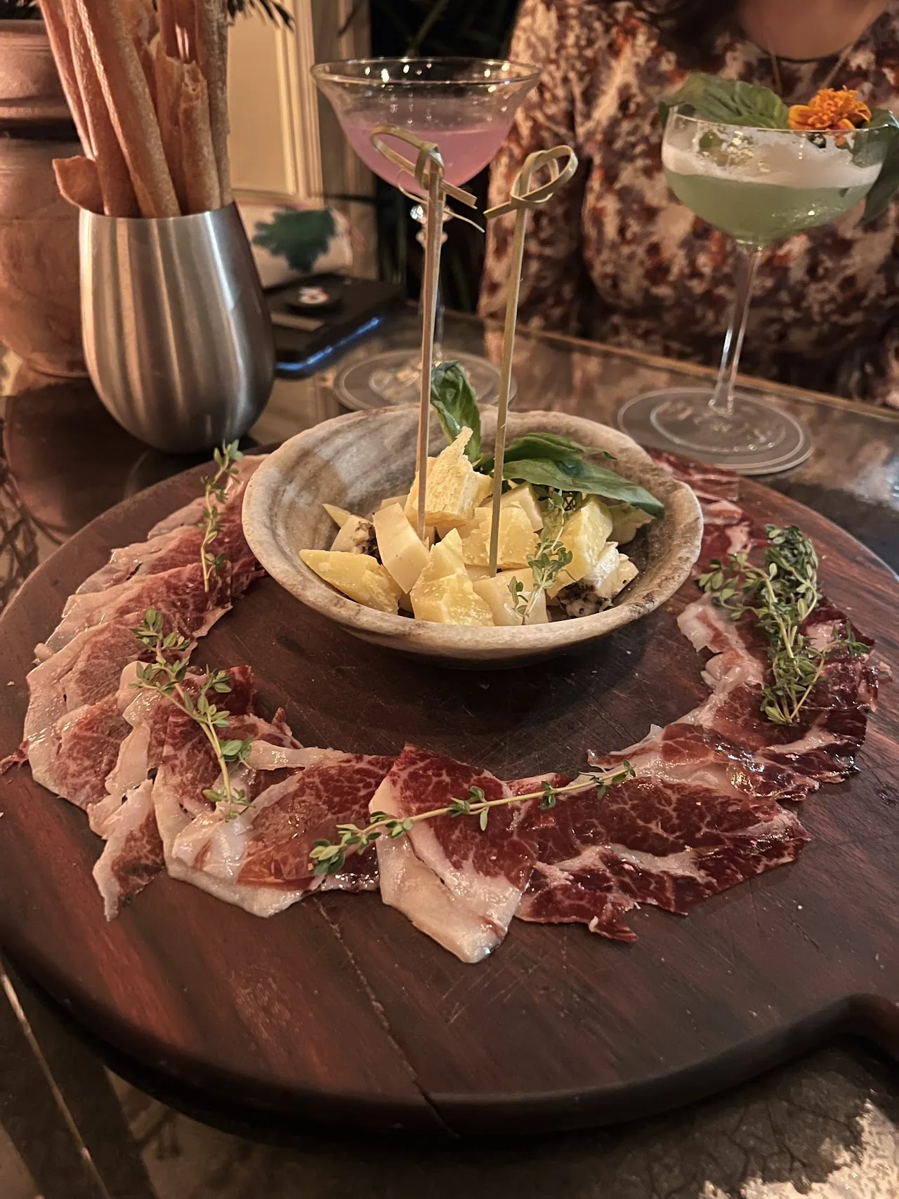 A charcuterie board with cured meats, cheese, and olives beside two cocktails