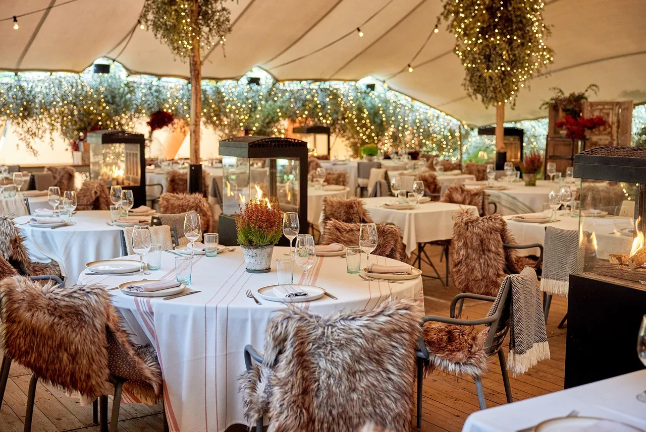 A tented garden dining area with round tables, string lights and fireplace heaters