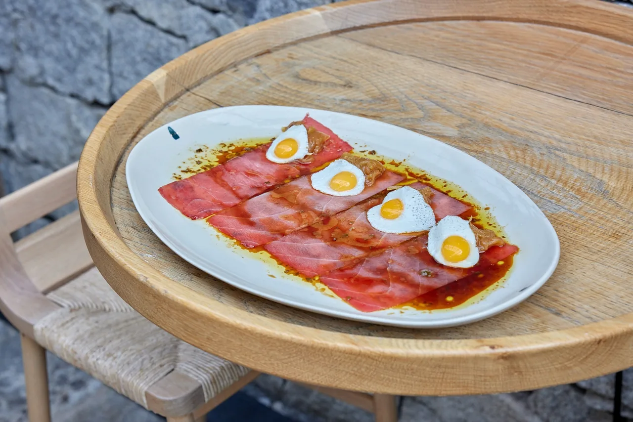 An oval plate of sliced cured meat topped with small fried eggs on a round wooden table
