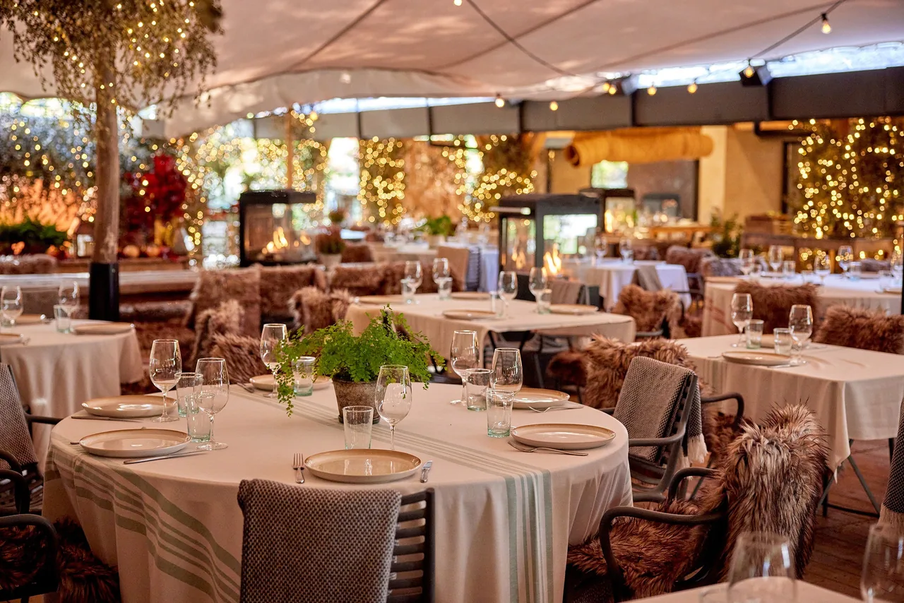 Round tables with white tablecloths and sheepskin-covered chairs under string lights on a covered garden terrace