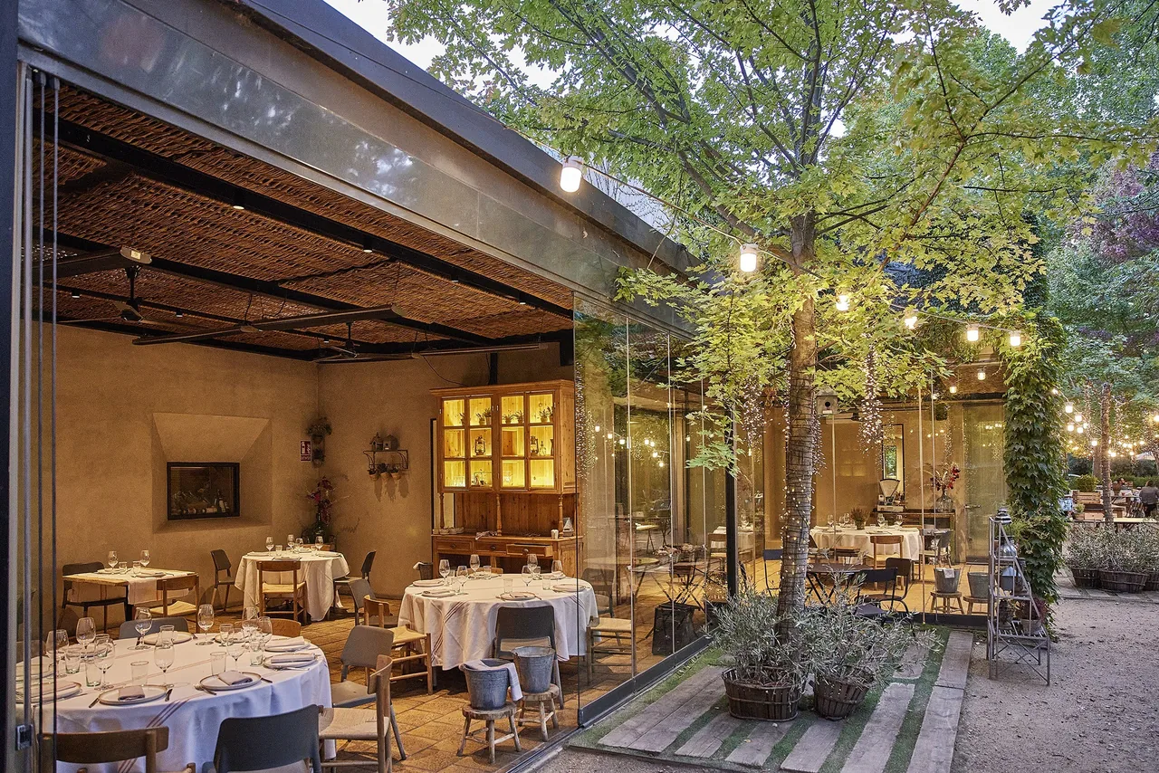 A glass-walled dining pavilion and laid tables under trees on a garden terrace at dusk