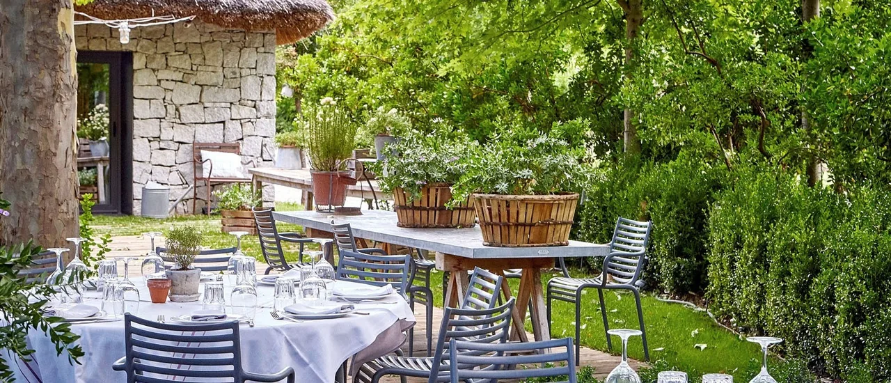 Tables laid with white cloths among trees and hedges on a garden terrace beside a stone building