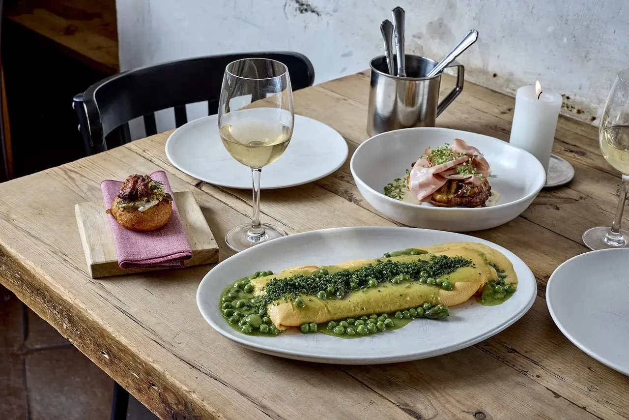 Table spread with a rolled omelette in pea puree, a bowl of ham-topped dish, a fried croquette and glasses of white wine