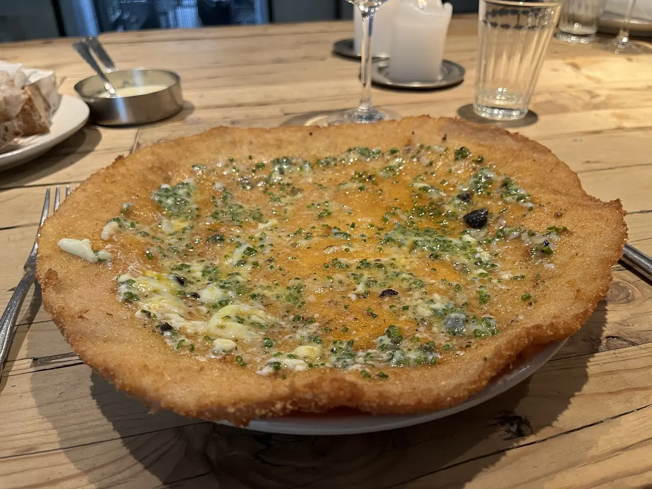 Large flat crispy dish topped with melted cheese, chives and black flecks, on a wood table with wine glass and candle
