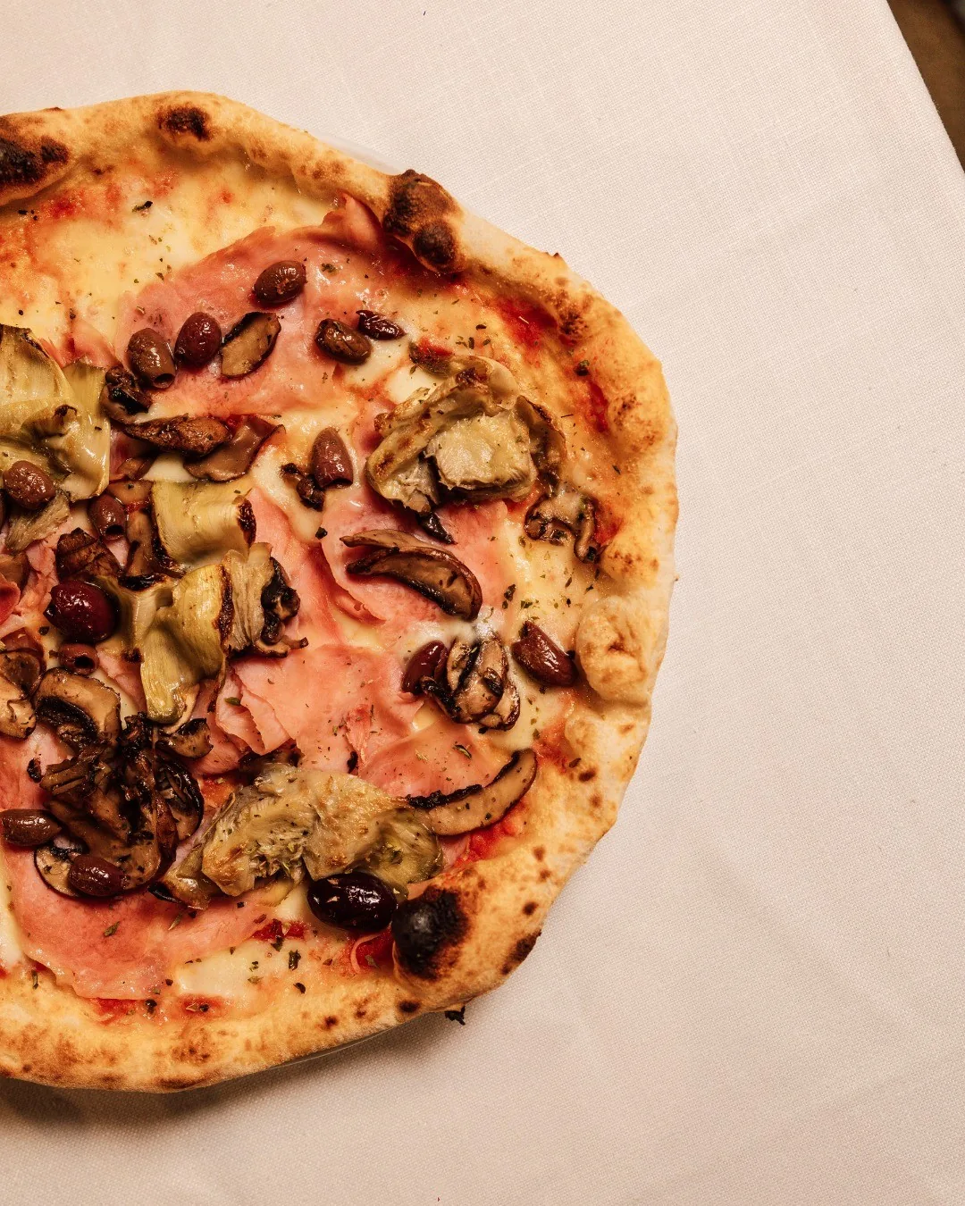 A round pizza topped with ham, mushrooms and artichokes on a white tablecloth