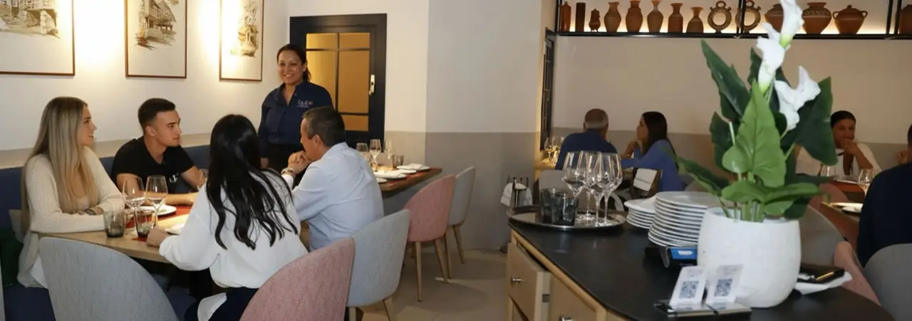Restaurant dining room with diners at set tables, framed watercolour prints, a shelf of terracotta jugs and a service station with stacked plates and wine glasses