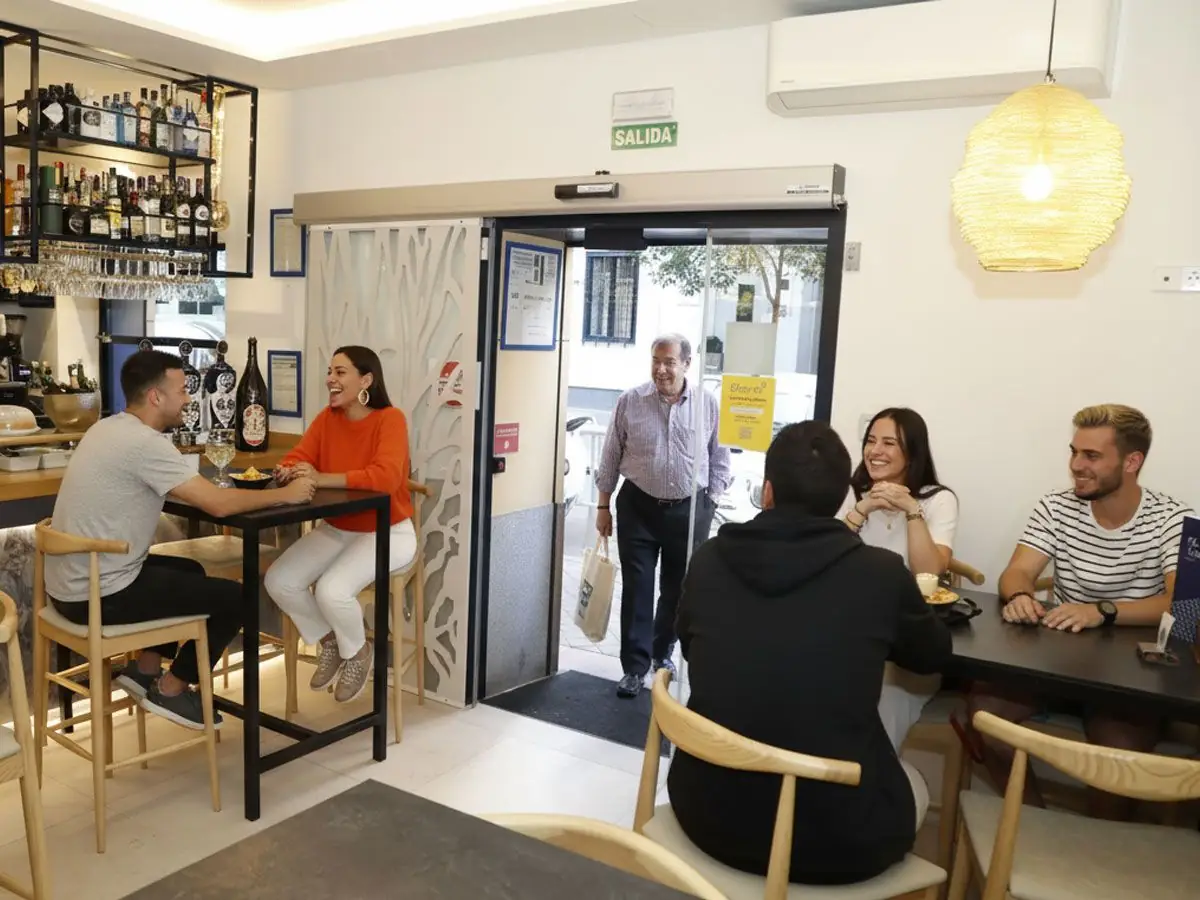 Bar and casual dining area with guests, back-bar spirits shelf, hanging glasses and glass sliding entrance door marked SALIDA