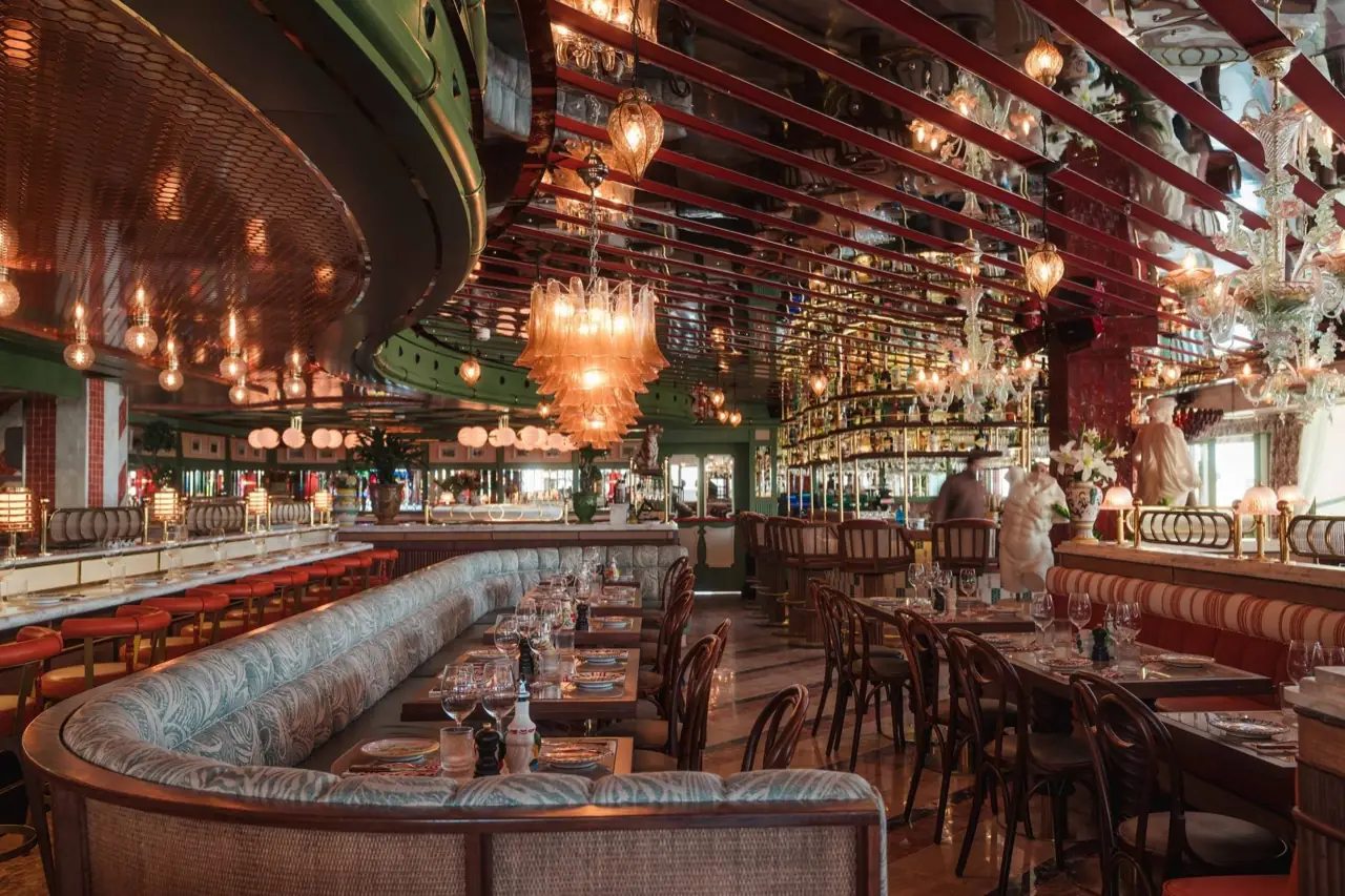 Opulent restaurant interior with a long curved pale-blue banquette, wooden bistro chairs at marble-topped tables, Murano glass chandeliers, mirrored ceiling and a lit back bar