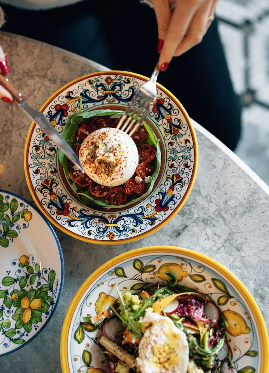 Patterned plates on a marble table with a round of white cheese, tomato and a colourful salad, seen from above