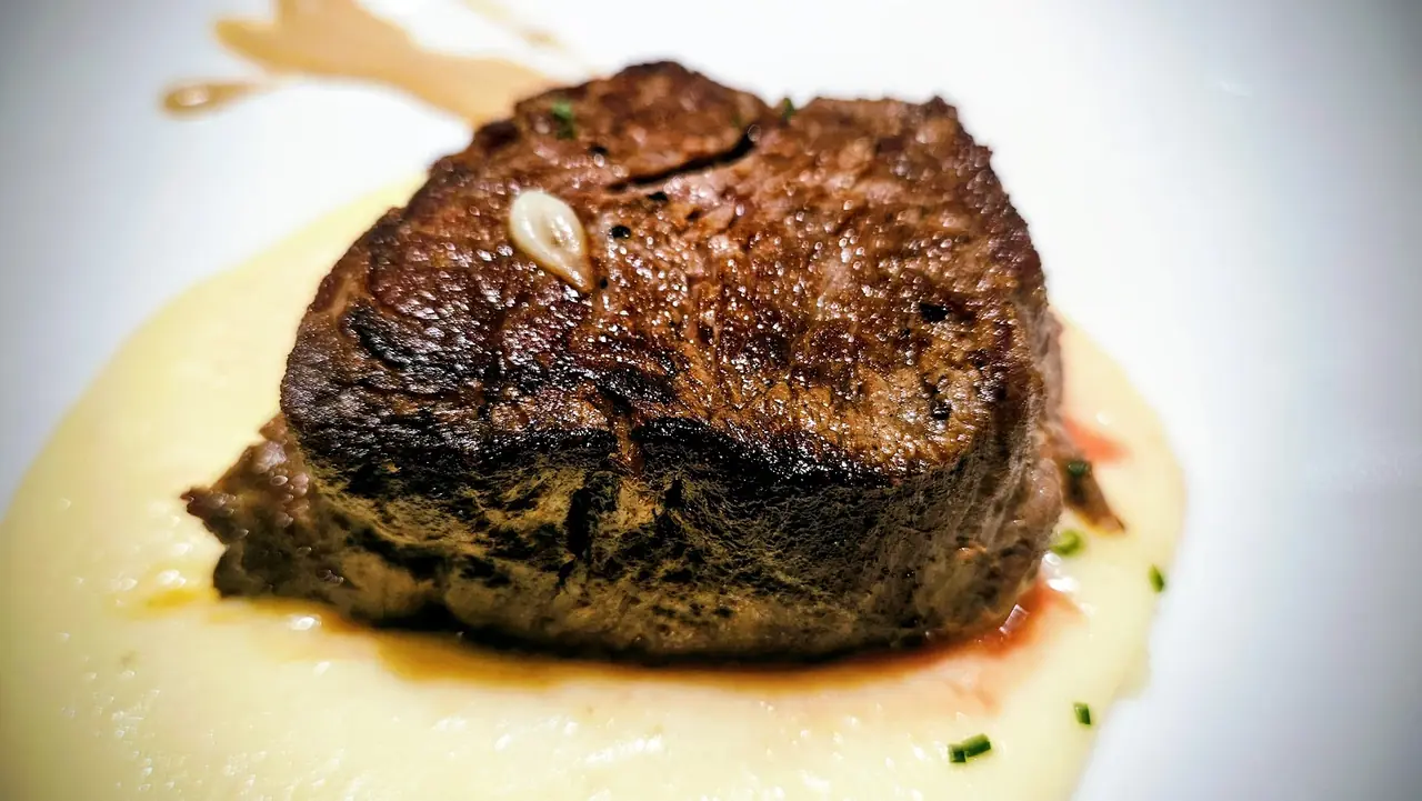 A seared steak with a garlic clove on top, plated over mashed potato with jus and chives