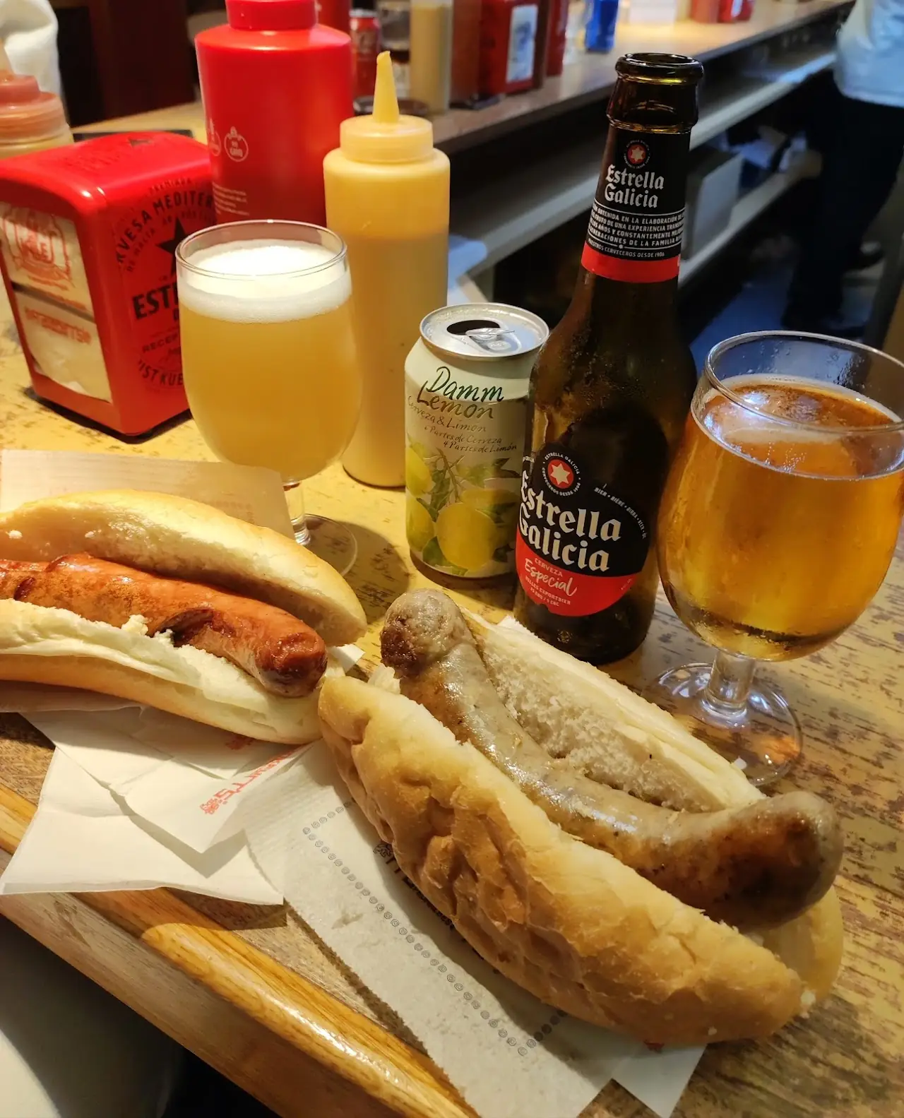 Hot dogs with condiments and glasses of beer on a wooden counter, at Frankfurt Pedralbes.