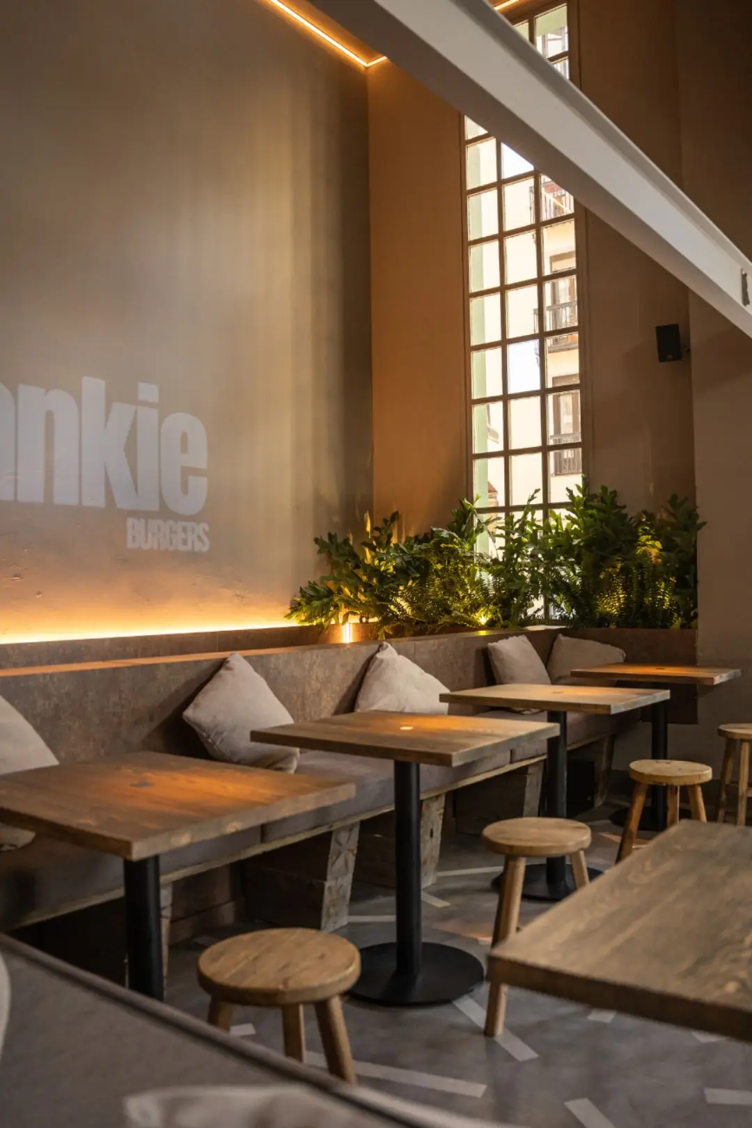 Interior of Frankie Burgers with a large white 'Frankie Burgers' logo on a taupe wall, banquette bench with grey cushions, wood tables and stools, and a tall grid window with plants