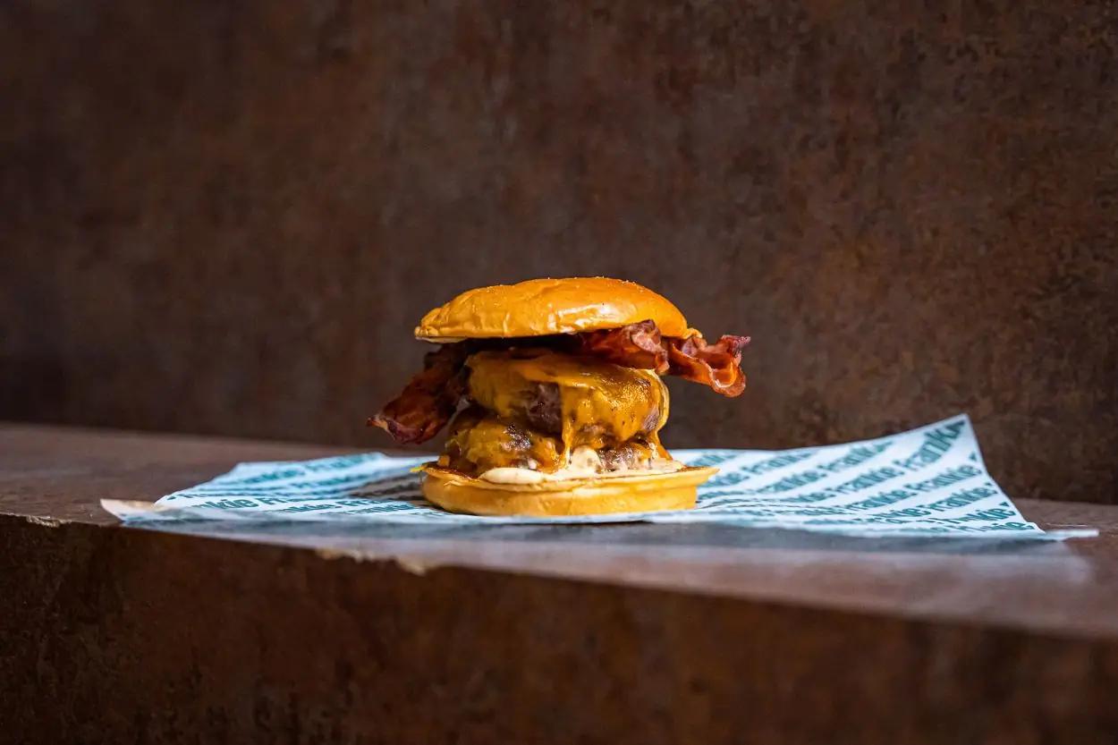 Bacon cheeseburger with melted cheese and crispy bacon on a brioche bun, sitting on branded 'Frankie' wax paper against a dark rust-coloured backdrop