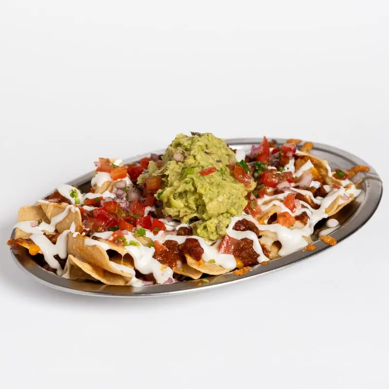 A platter of tortilla chip nachos topped with guacamole, pico de tomato, and a drizzle of white sauce.