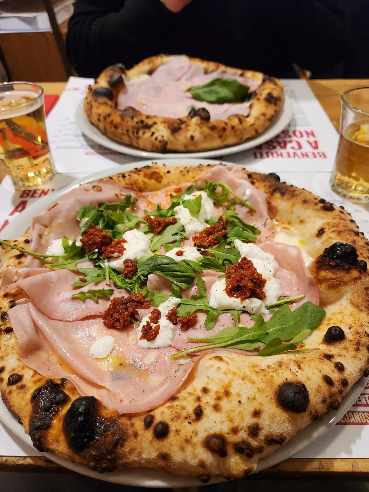 Two pizzas topped with ham, rocket and sun-dried tomatoes on plates, with a glass of beer beside them