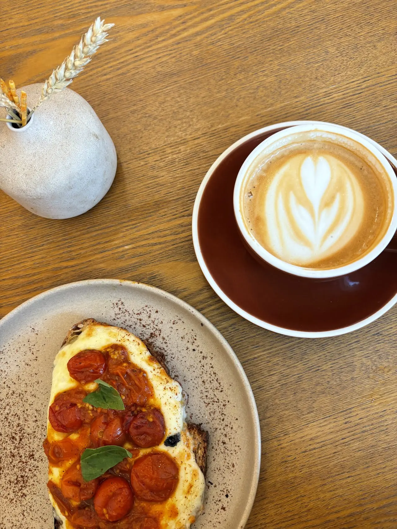 Toast topped with roasted cherry tomatoes and basil beside a latte with leaf latte art, on a wooden table