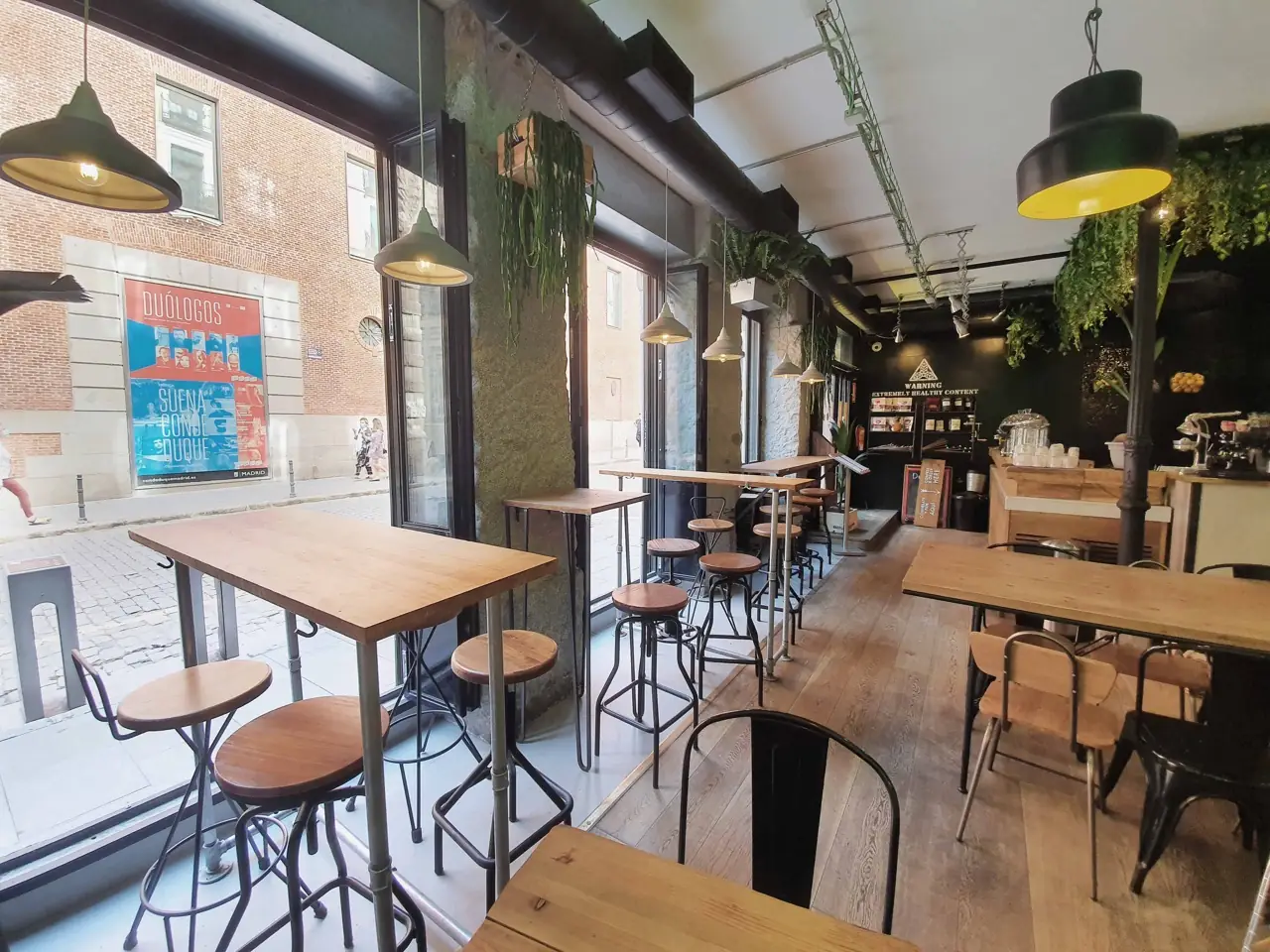Interior of a plant-filled cafe with reclaimed-wood high tables, metal bar stools, industrial pendant lamps, brick walls, a coffee bar at the back and a street view through the glass front