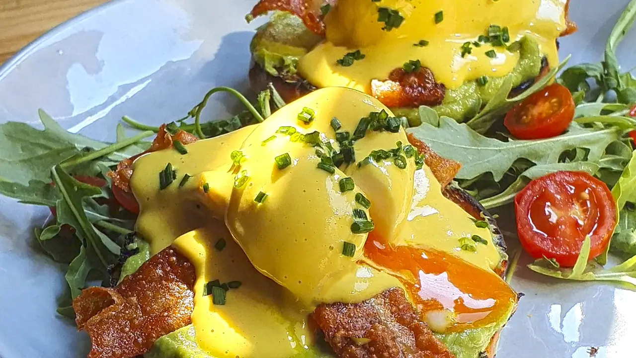 Close-up of eggs benedict with poached eggs, yellow hollandaise, chopped chives, crispy bacon and arugula-and-cherry-tomato salad on a grey plate